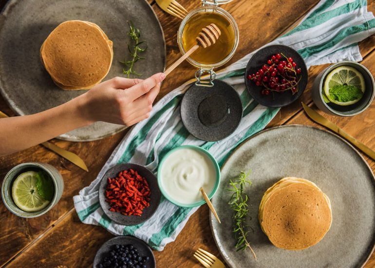 Widok z góry na śniadanie z naleśnikami, owocami, jogurtem i miodem na tle drewnianego designerskiego stołu