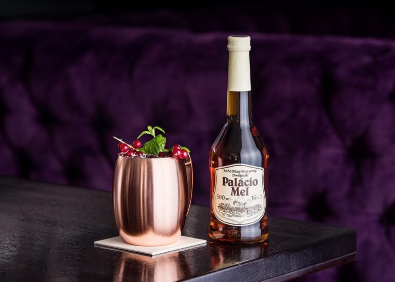 Butelka miodu pitnego Palacio Mel z drinkiem w miedzianym kubku na tle fioletowej kanapy w luksusowej restauracji