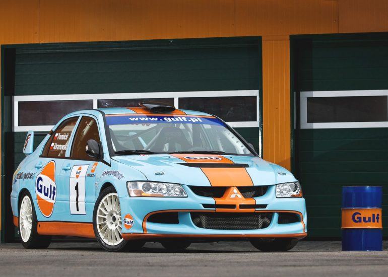 Samochód rajdowy Mitsubishi Lancer Evo VIII w barwach Gulf na tle garażu z beczką oleju silnikowego