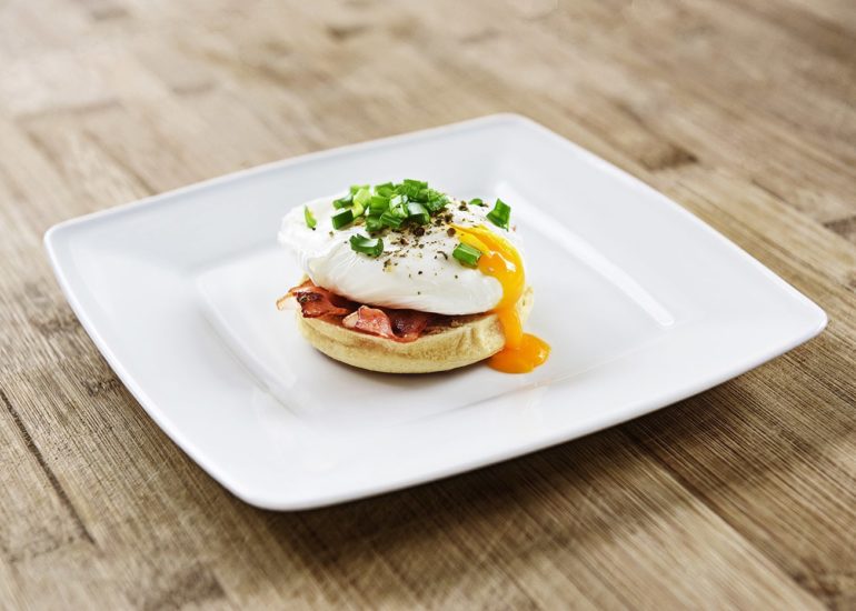 Muffin angielski z jajkiem po benedyktyńsku, smażonym bekonem, szczypiorkiem i pieprzem na białym talerzu
